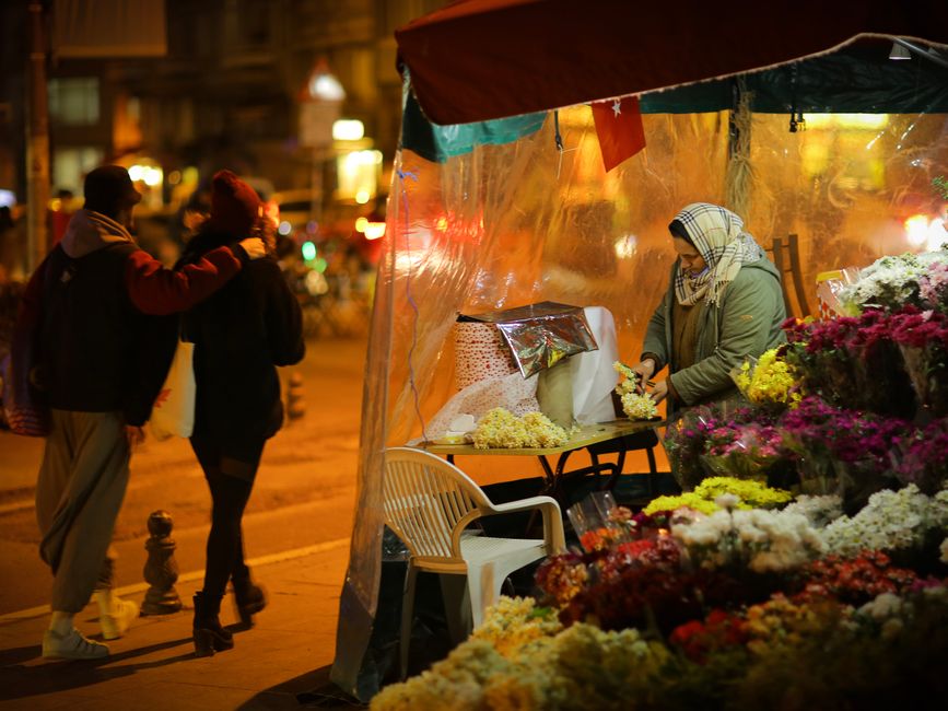 A flower vendor at a street stall during evening hours, arranging and displaying a vibrant assortment of fresh flowers including roses, chrysanthemums, and lilies in shades of white, yellow, pink, and
