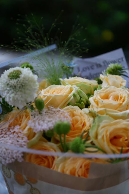 Close-up of a floral bouquet featuring pale peach roses with soft, velvety petals, surrounded by delicate white and green filler flowers, including small white pom-pom-like blooms and green buds. The 