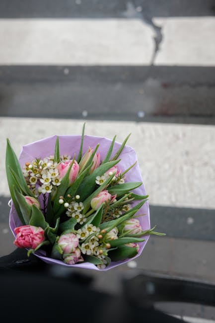 A close-up of a bouquet of fresh flowers held by a person, featuring pink tulips, small white daisy-like flowers, and green foliage wrapped in light purple paper. The arrangement sits against a blurre