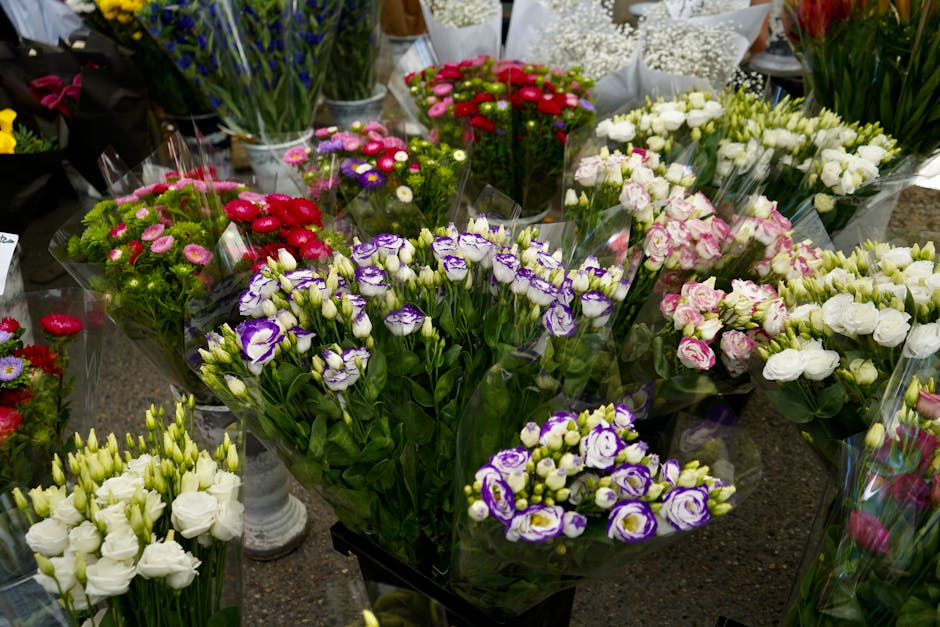 A variety of fresh floral bouquets arranged on a display table in a flower shop, featuring lisianthus in purple and white, pink carnations, white roses, and other colorful blooms. The flowers are wrap